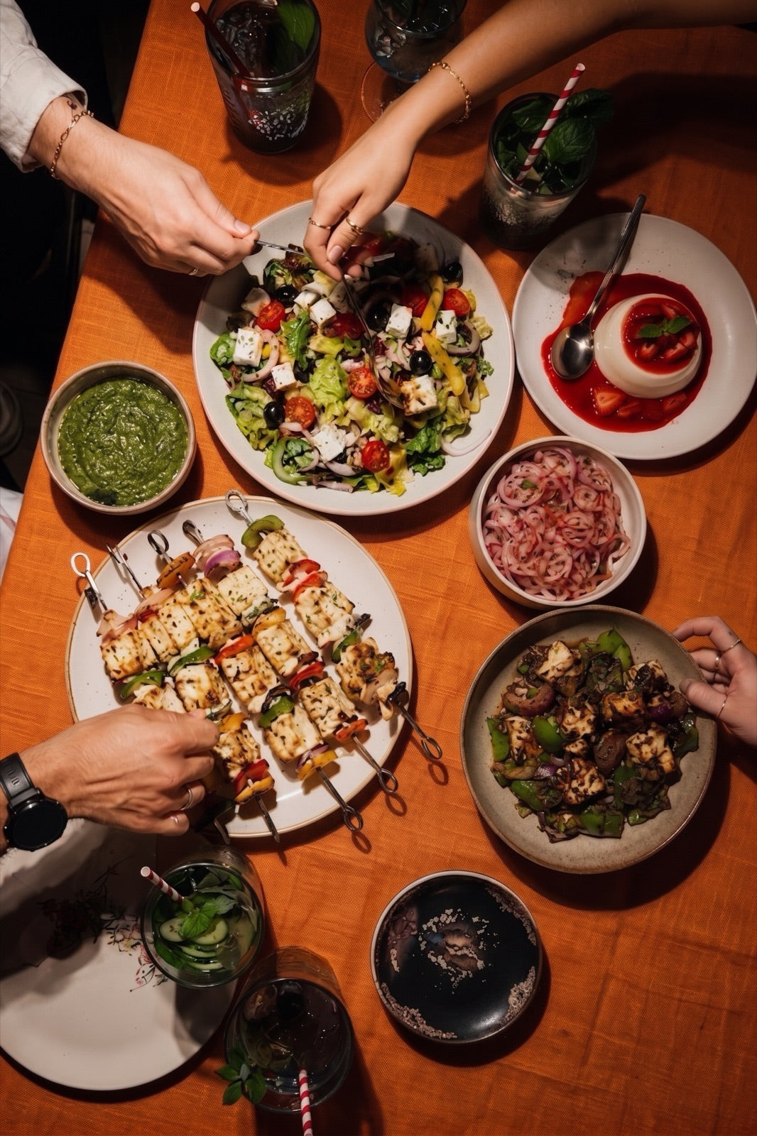 Overhead shot of hands sharing a spread of grilled paneer skewers, Greek salad, pickled onions, and panna cotta at the table