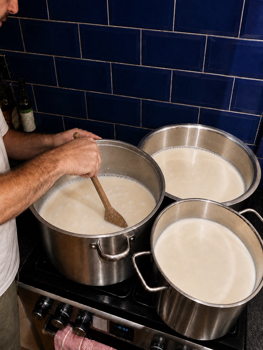 Hand stirring fresh milk with a wooden spatula in a large steel pot on the stove