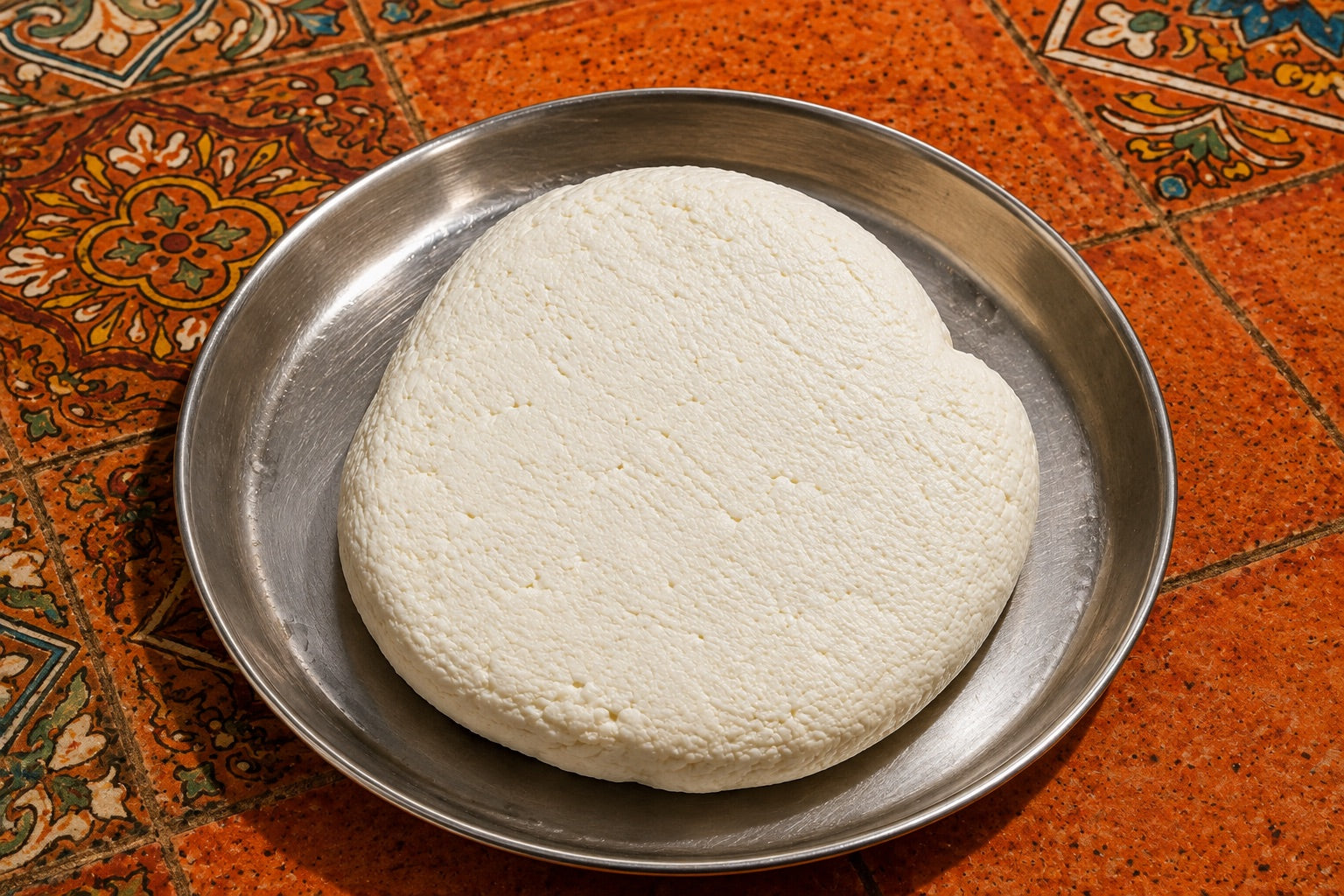 A freshly pressed round of paneer resting on a steel plate on hand-painted tiles